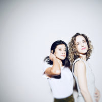 Two women standing against a light grey background in light coloured tops
