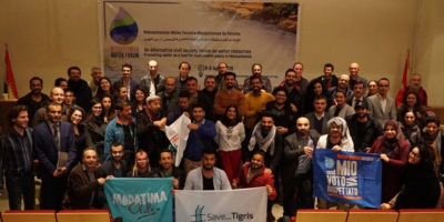 Over 50 people gathered in a room in front of a banner for the Mesopotamian Water Forum, the ones in front holding placards which read 