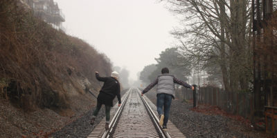 Two people photographed from the back walking on the left and right rails of traintracks in the country.