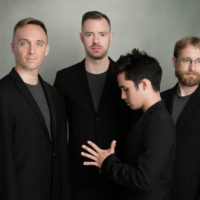 Four men posing in dark suits, one holding his hand out with his palm facing toward him