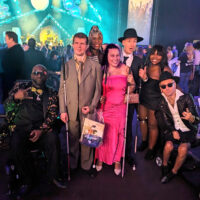 Photo provided by Recording Artists and Musicians with Disabilities (RAMPD) -Seven artists pose together for a photo at the 2024 Grammy afterparty. They are smiling and dressed in stylish attire. The background features a brightly lit stage, elegantly decorated venue with colorful lighting and a crowd of people in the distance.